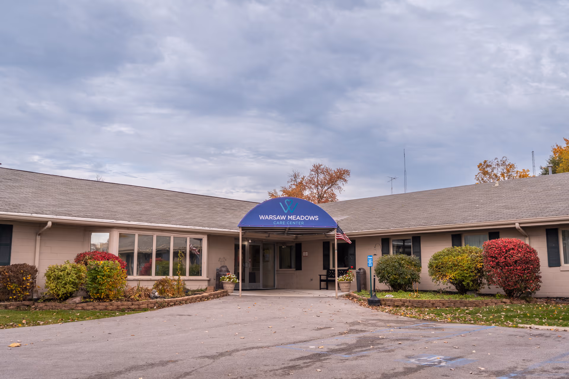Exterior front view of Warsaw Meadows Care Center, a single-story building with a gray roof and beige walls. The entrance has a blue awning with the facility's name and logo. There are bushes and small trees with autumn foliage around the building, and an American flag is displayed near the entrance.