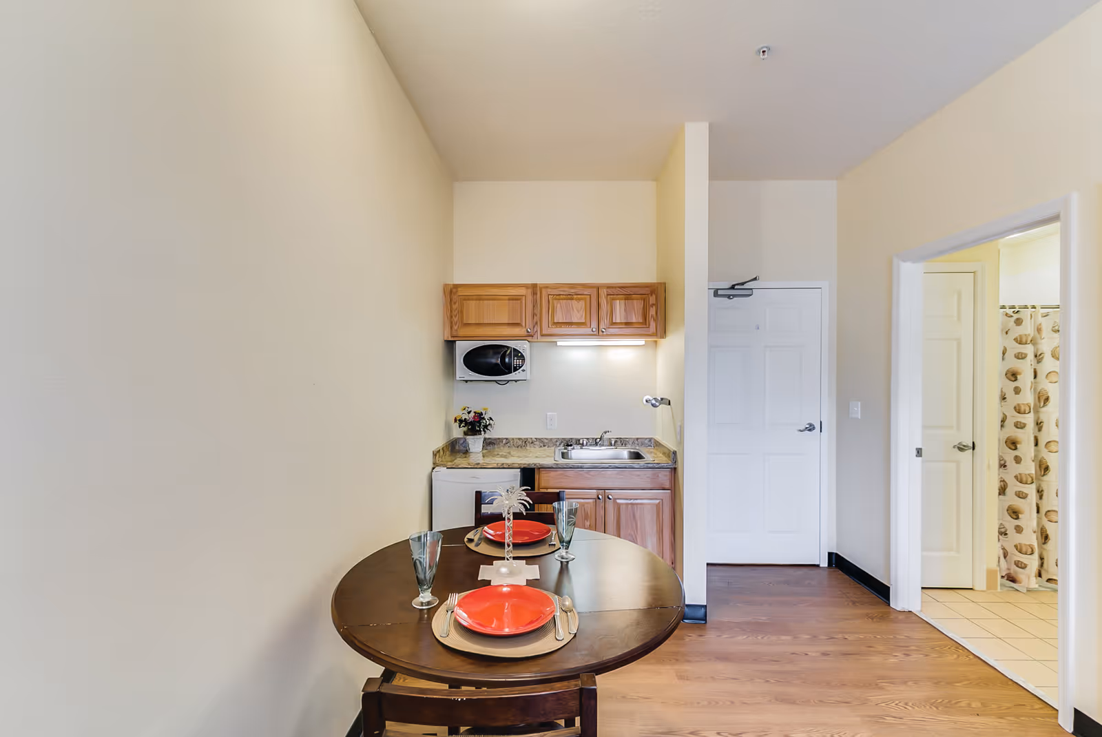 Small kitchenette area with wooden cabinets, a microwave, a sink, and a mini refrigerator. In front of the kitchenette is a round wooden dining table set for two with red plates, silverware, and glasses. To the right, there is a doorway leading to a bathroom with a shower curtain visible.
