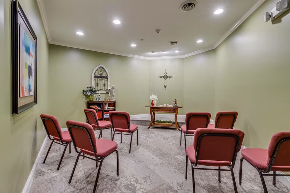 Small chapel-style room with green walls and red padded chairs arranged facing a wooden altar and wall-mounted cross.