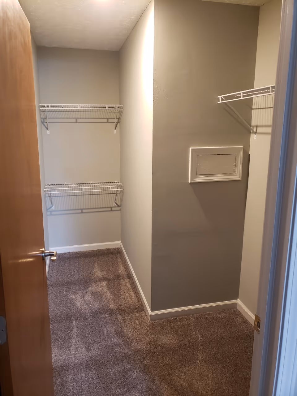 Small empty closet with beige walls and brown carpet. There are white wire shelves mounted on the walls and a wooden door partially open.