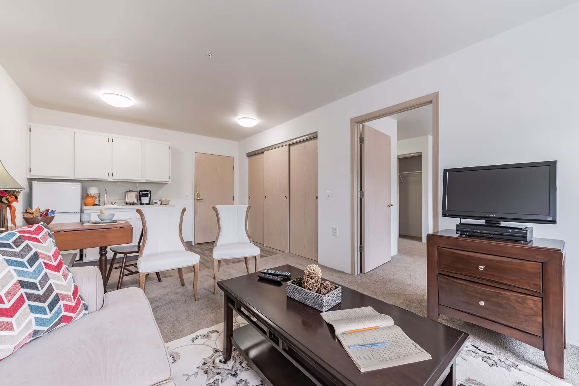 Bright furnished living room with a sofa, coffee table and TV, two dining chairs and a small kitchenette visible in the background.