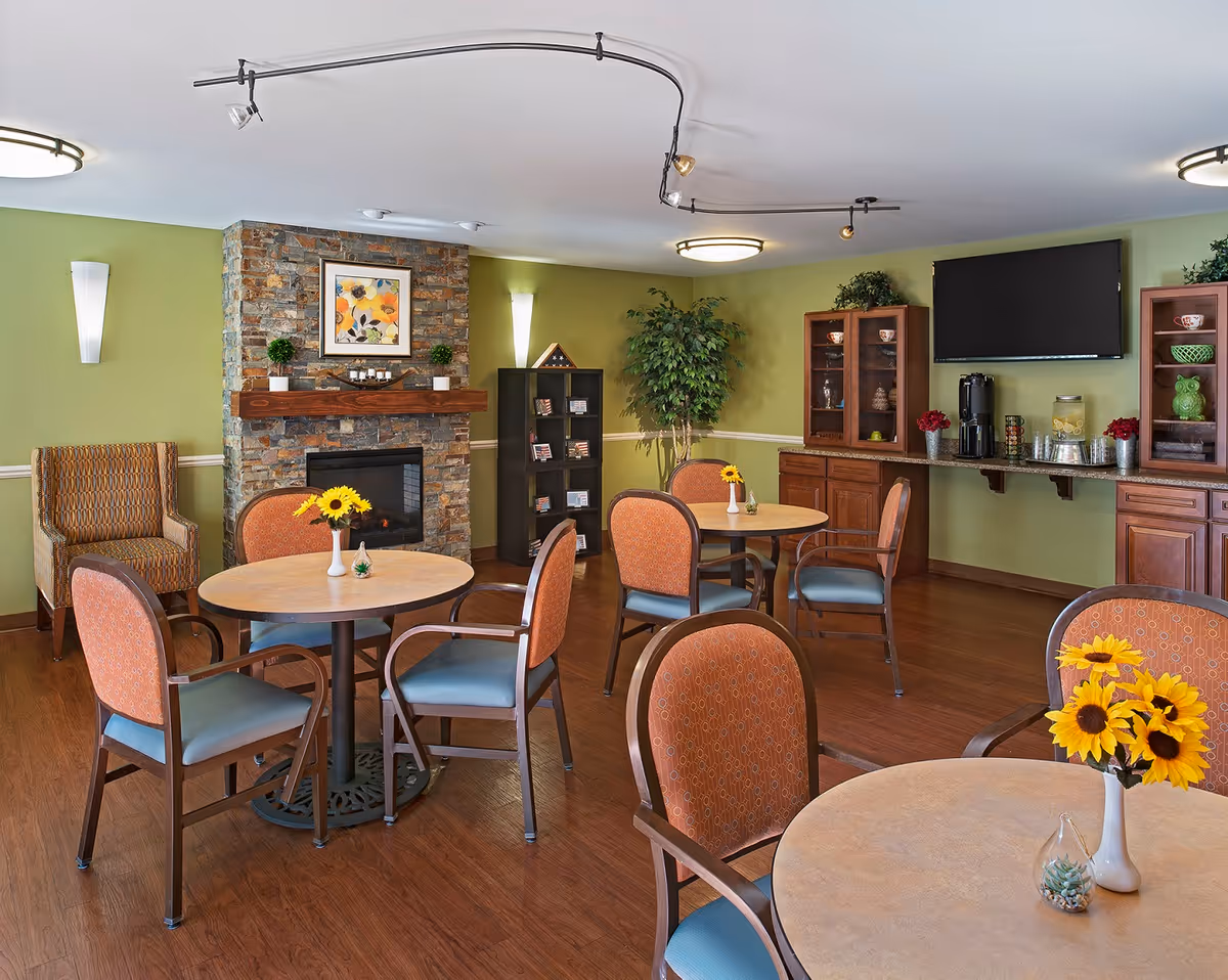 A cozy common area with round tables and chairs arranged for seating. The room features a stone fireplace with a framed floral artwork above it, green walls, wooden flooring, and a cabinet with a coffee station and a mounted flat-screen TV. There are decorative plants and vases with sunflowers on the tables.