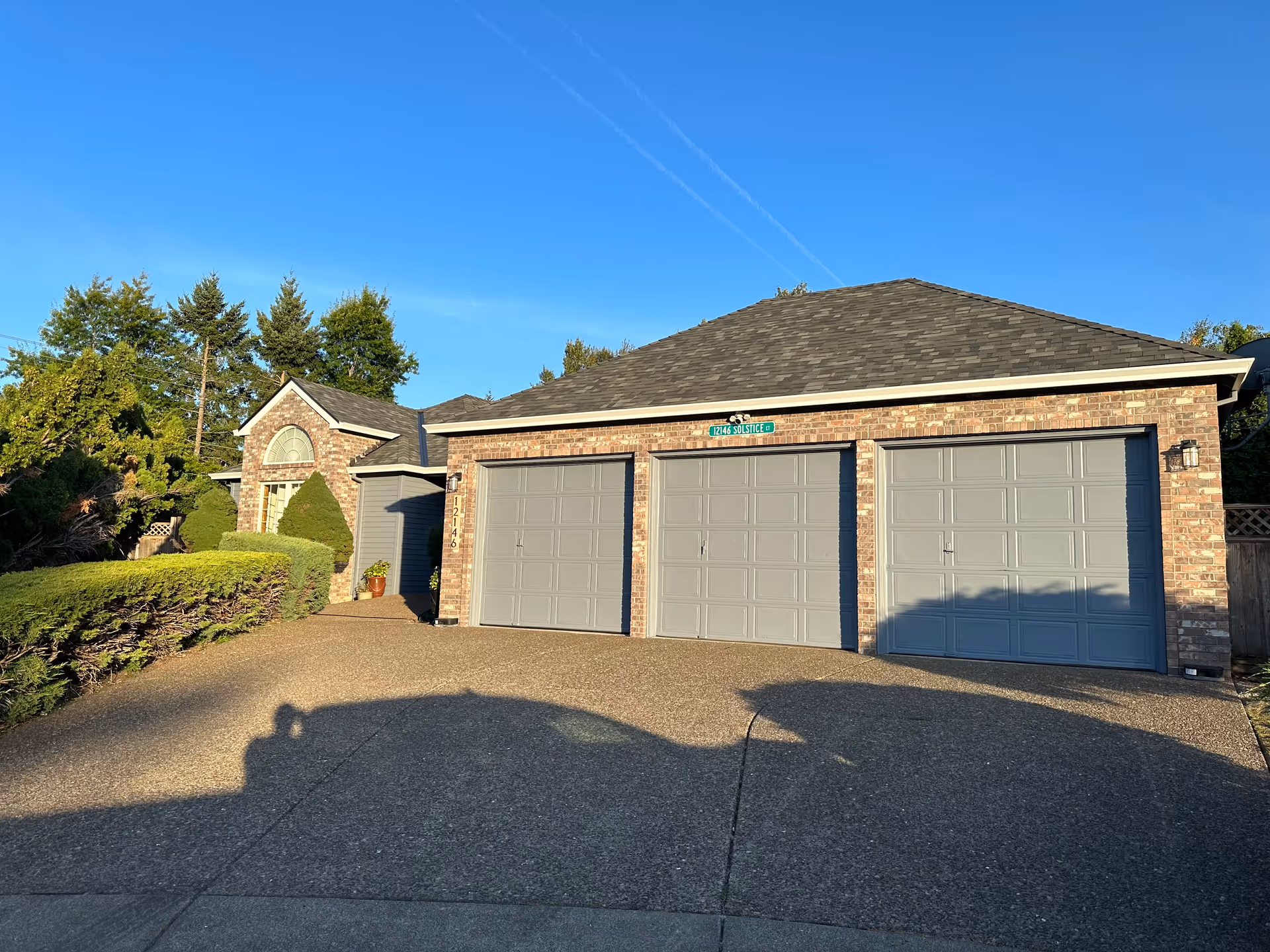 Exterior view of a brick building with three gray garage doors and a driveway in front. The building is surrounded by green trees and bushes under a clear blue sky.