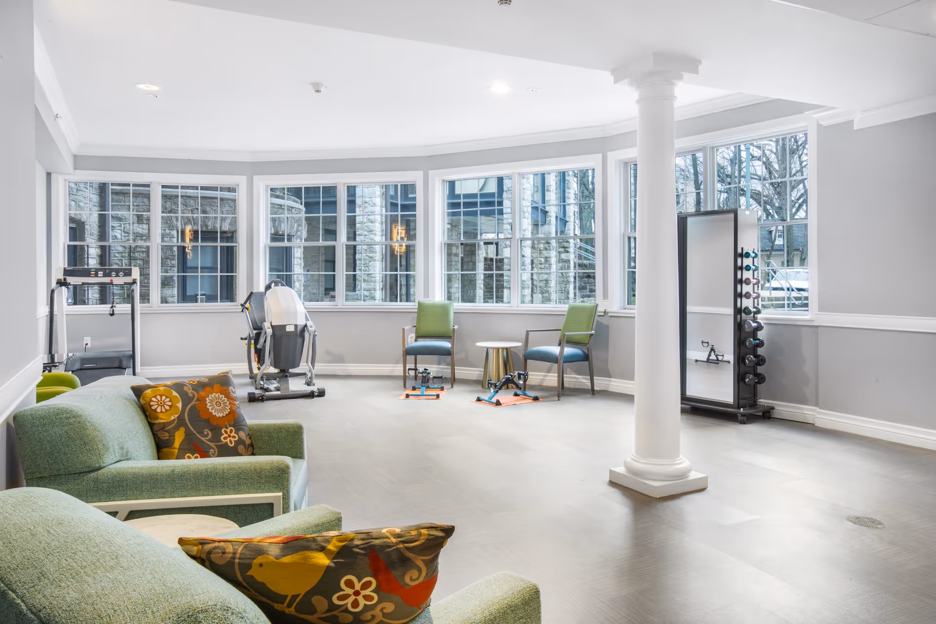 A bright and spacious exercise room with large windows letting in natural light. The room contains two green armchairs with colorful patterned pillows, a small round table between two chairs, exercise equipment including a stationary bike, hand weights on a rack, and pedal exercisers on the floor. The walls are painted light gray with white trim, and there is a white column in the center of the room.