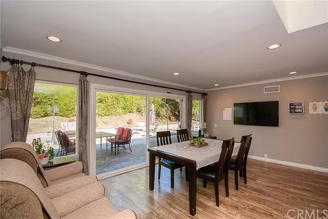 Interior view of a senior living facility dining area with a wooden table covered by a white lace tablecloth and six chairs. There is a beige sofa on the left side and large sliding glass doors leading to an outdoor patio with chairs and greenery. A flat-screen TV is mounted on the wall to the right.