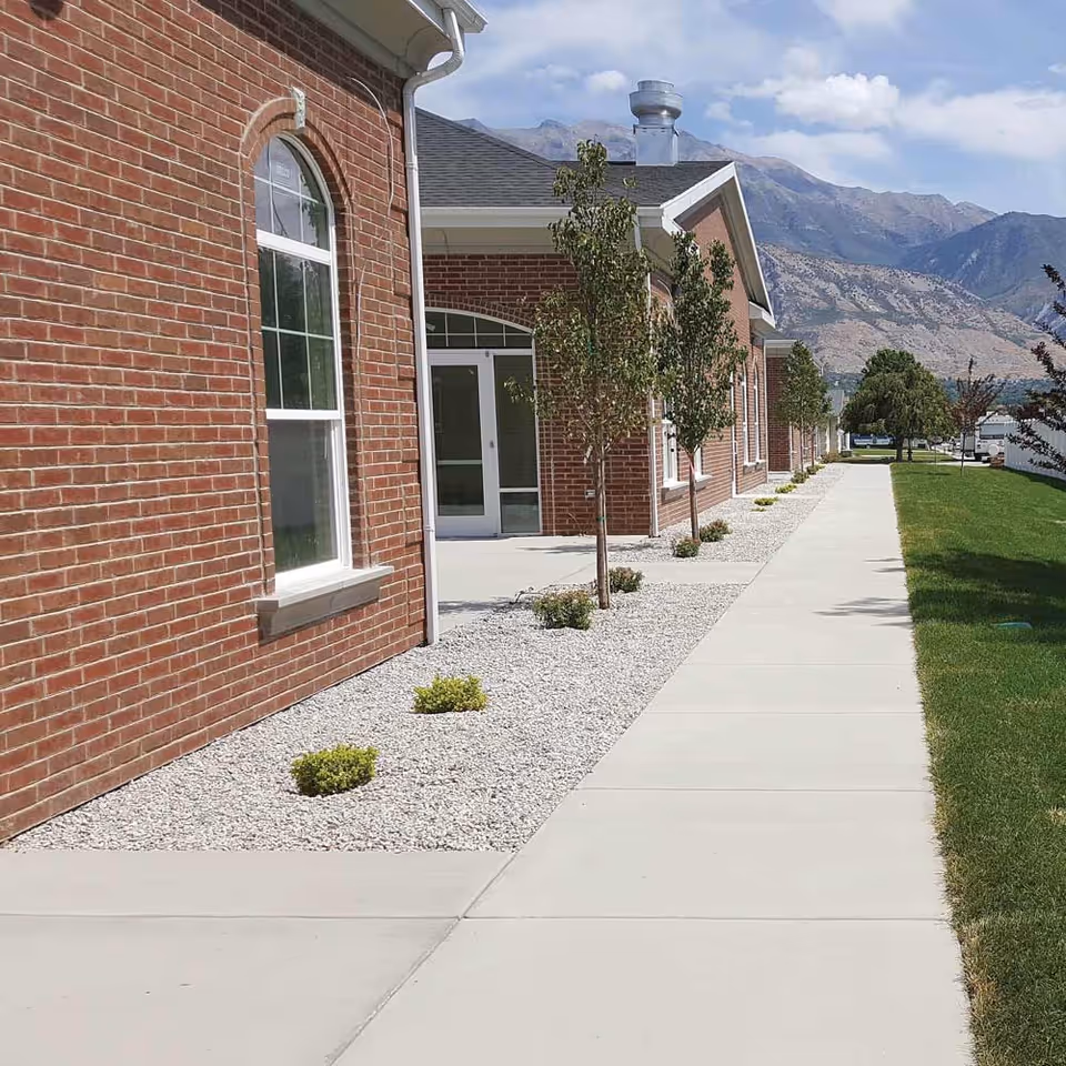 Front exterior of a red-brick assisted living building with a concrete sidewalk, small trees and gravel landscaping, and mountains in the background.