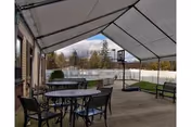 Outdoor covered patio area with a round metal table and several chairs. There is a basketball hoop on the concrete floor and a white fence enclosing the grassy yard. Trees and a cloudy sky are visible in the background.