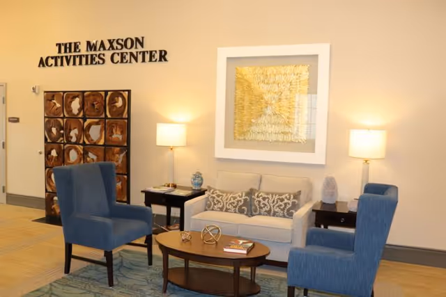 Seating area in an activities center with a sofa, two blue armchairs, a coffee table, lamps, wall art, and a sign reading "THE MAXSON ACTIVITIES CENTER".