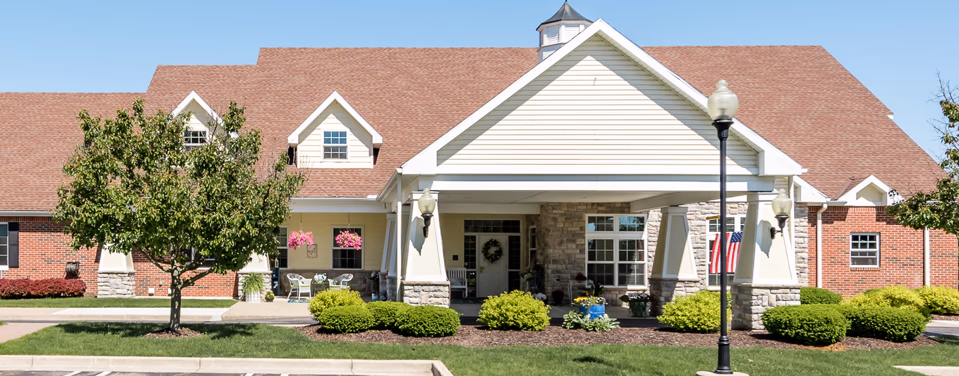 Front entrance of a single-story senior living building with a covered portico, landscaping, and an American flag.