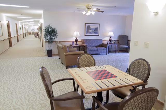 A well-lit common area in a senior living facility featuring a table with a checkerboard and backgammon game design surrounded by four chairs. In the background, there is a seating area with a sofa, two armchairs, two table lamps, a framed landscape painting on the wall, and a potted plant. A long hallway extends to the left with handrails and framed pictures on the walls.