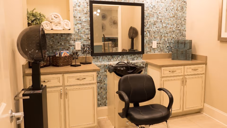 Interior view of a salon area in an assisted living facility featuring a black salon chair in front of a black hair washing sink. The background has a mosaic tile wall with a large rectangular mirror mounted on it. There are cream-colored cabinets with drawers and decorative items, including rolled white towels, a plant, and small storage boxes on the countertops.