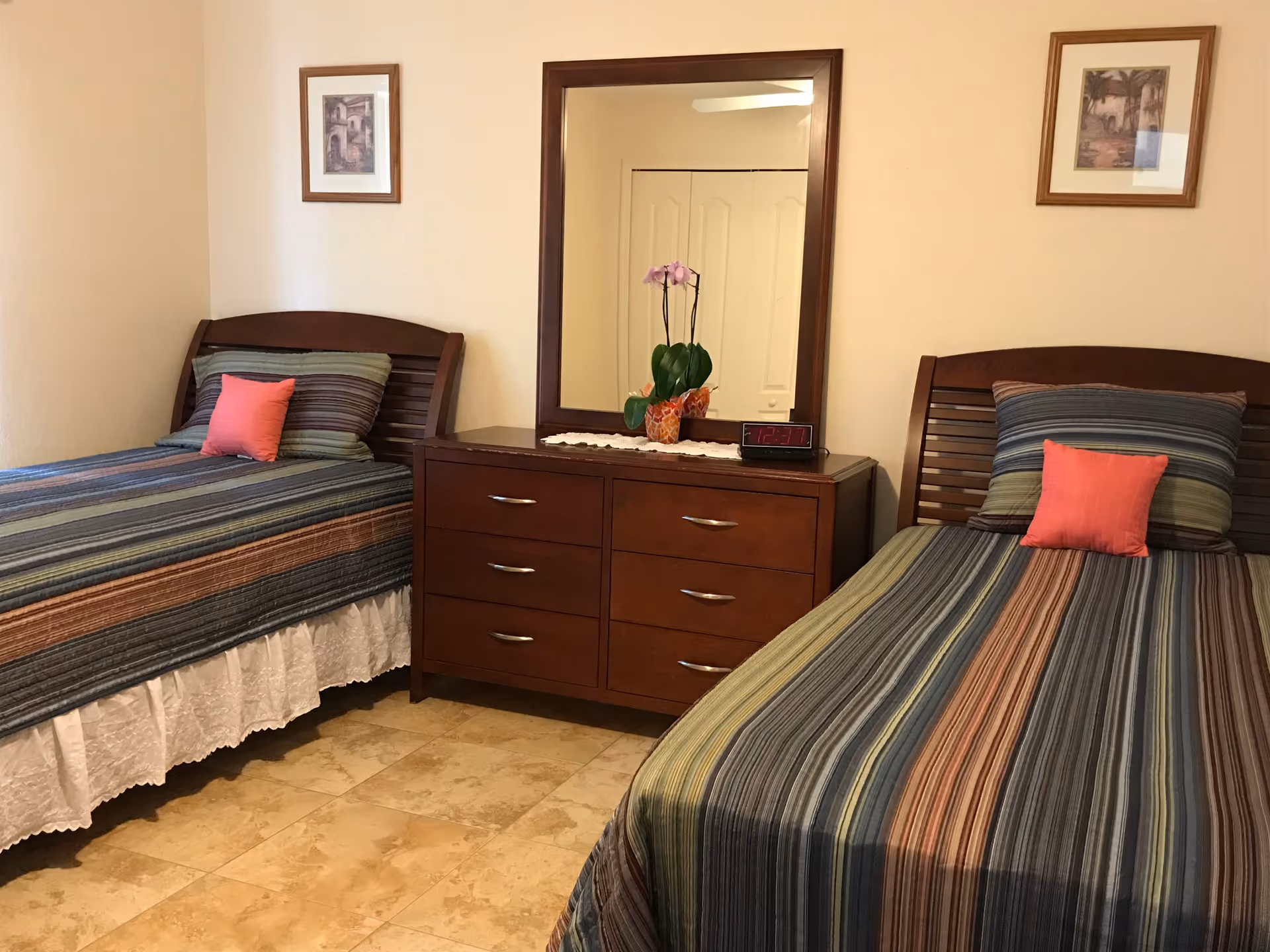 A tidy bedroom with two twin beds with striped bedding, a central wooden dresser with mirror, and decorative pillows and artwork on the walls.