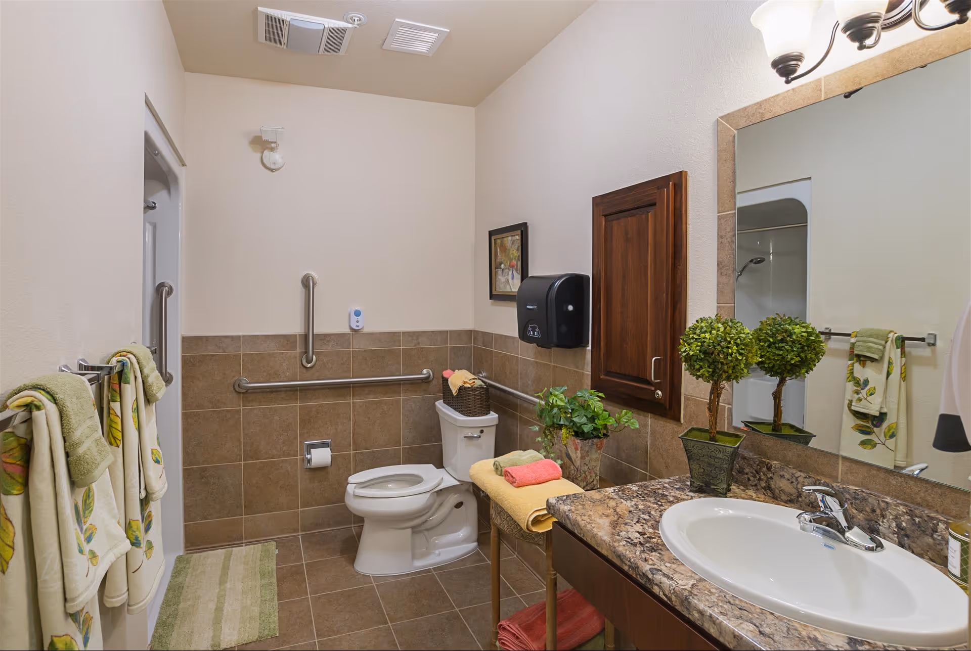 A clean and accessible bathroom with beige tiled walls and floor. The bathroom features a white toilet with grab bars on the walls around it, a granite countertop with a white sink, a large mirror, a wooden cabinet, and decorative plants. Towels with leaf patterns hang on a towel rack, and there is a small bench with folded towels and a basket on top.