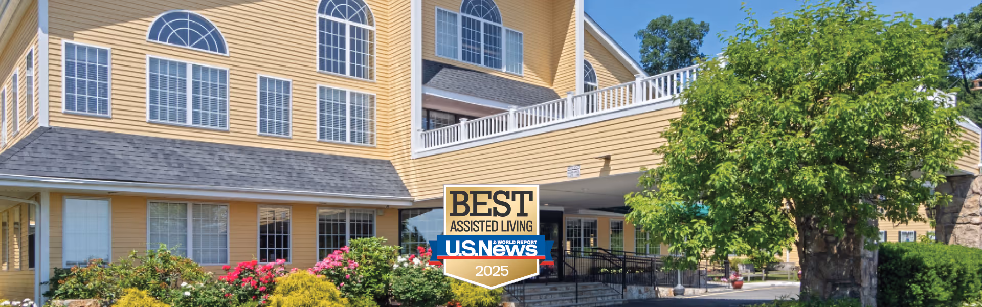 Exterior view of a yellow multi-story assisted living facility with large windows and a covered entrance. There are green bushes and flowering plants in front of the building, and a tree to the right. A sign in front reads 'Best Assisted Living U.S. News 2025'.