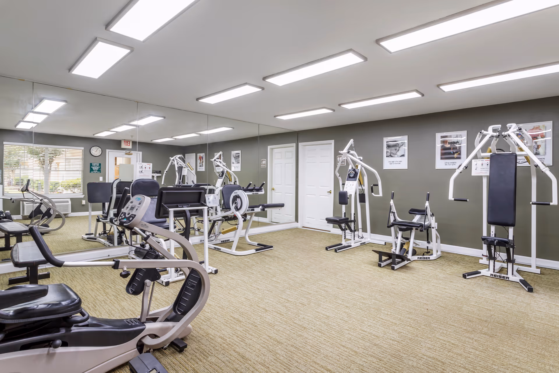 A well-lit fitness room with various exercise machines including a recumbent bike, weight machines, and benches. The room has a large mirror covering one wall, beige carpet flooring, and gray walls with framed motivational posters. A window with blinds and an exit door are visible in the background.