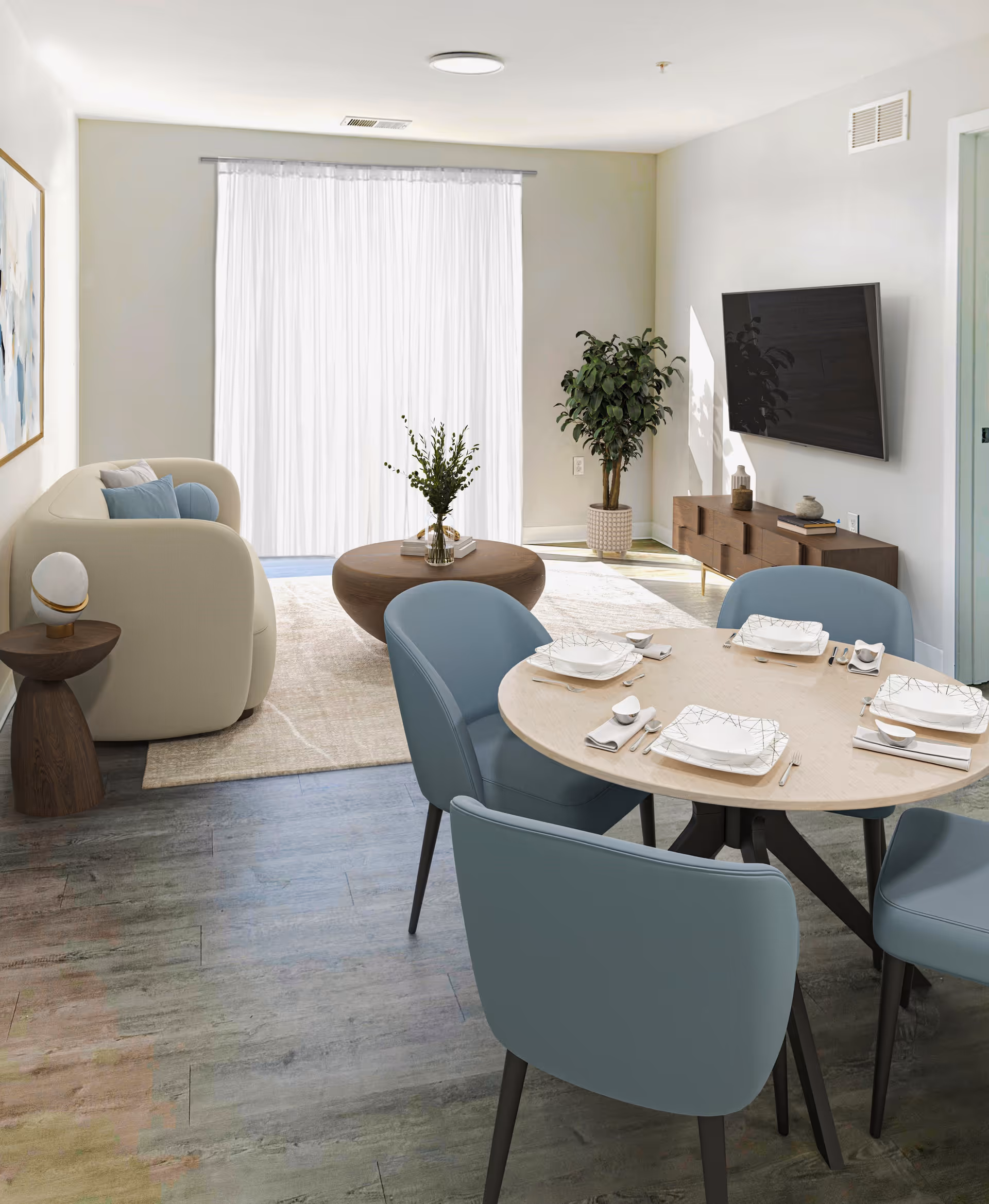 A modern living and dining area with a round dining table set for four with blue chairs. The living area has a beige sofa, a round wooden coffee table with a vase of greenery, a side table with a lamp, a large flat-screen TV mounted on the wall, and a potted plant in the corner. Light filters through sheer white curtains covering a large window or sliding door.