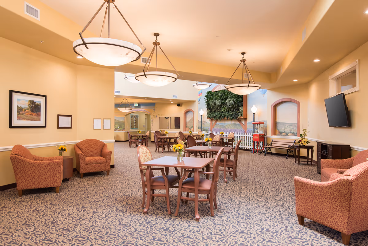 A spacious and well-lit common area in a senior living facility with multiple tables and chairs arranged for dining or socializing. The room features warm yellow walls, patterned carpet, large hanging light fixtures, and comfortable armchairs. There are framed pictures on the walls, a flat-screen TV mounted on the right wall, and a decorative green plant wall with a white picket fence in the background. A popcorn machine and benches are also visible near the back wall.