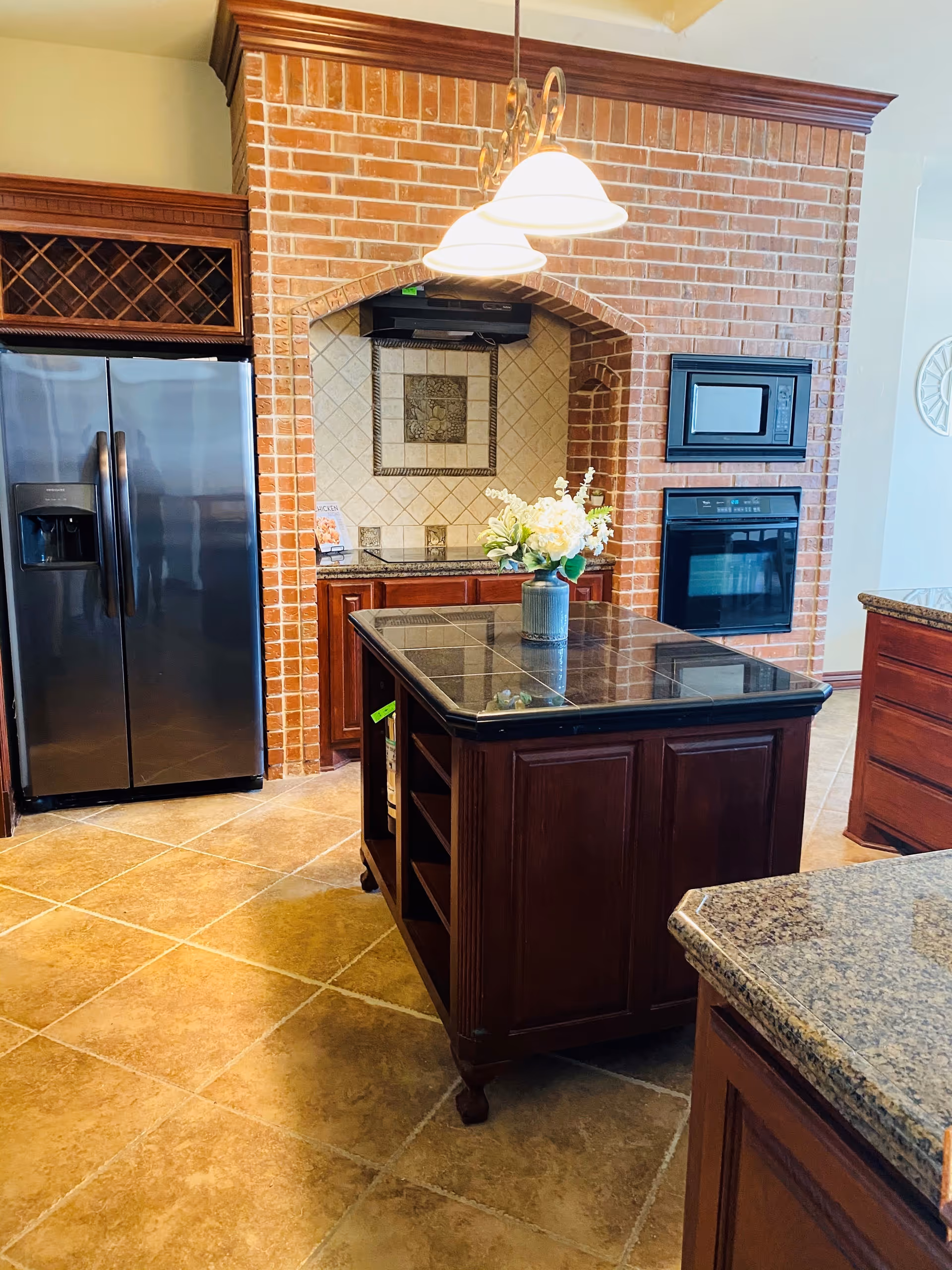 A kitchen with a central island topped with black tiles and a vase of flowers. The kitchen features a stainless steel refrigerator, a built-in microwave, and an oven set into a brick archway with tiled backsplash. The floor is tiled in a warm beige color and the cabinetry is dark wood.