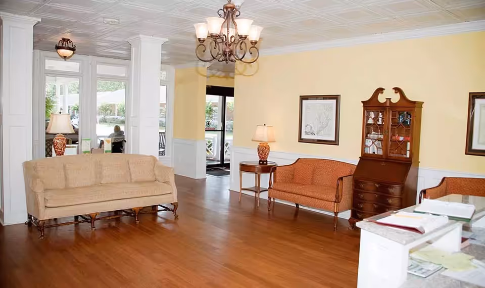 A bright and spacious living room area with wooden flooring, beige and orange upholstered sofas, a wooden cabinet with glass doors, framed artwork on the yellow walls, a chandelier hanging from the ceiling, and table lamps on side tables. Large windows and a glass door provide a view of the outdoor area.