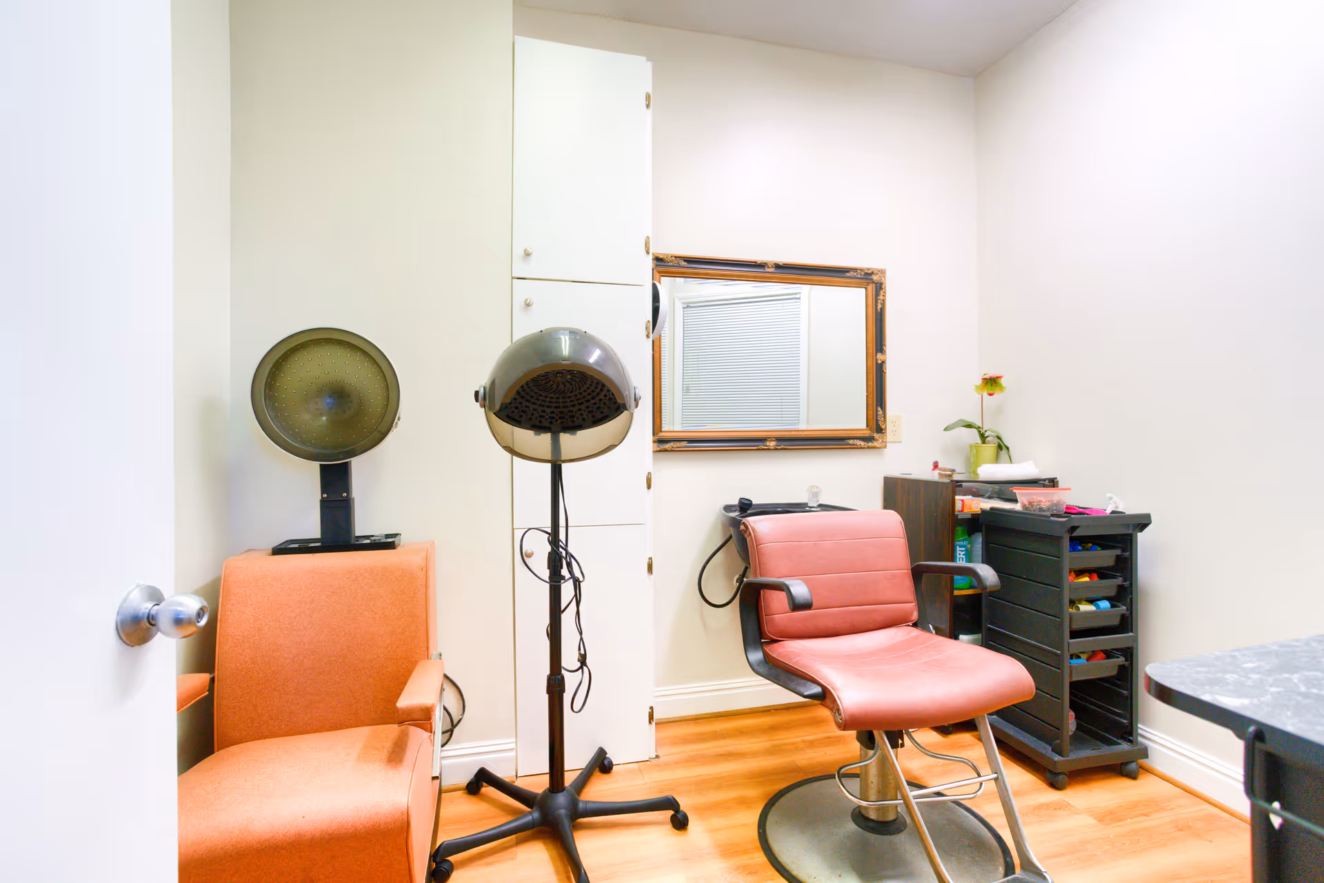 A small salon or beauty room with a red salon chair, a hair dryer on a stand, an orange armchair, a mirror on the wall, a small black rolling cart with various supplies, and a small sink. The room has light-colored walls and wooden flooring.