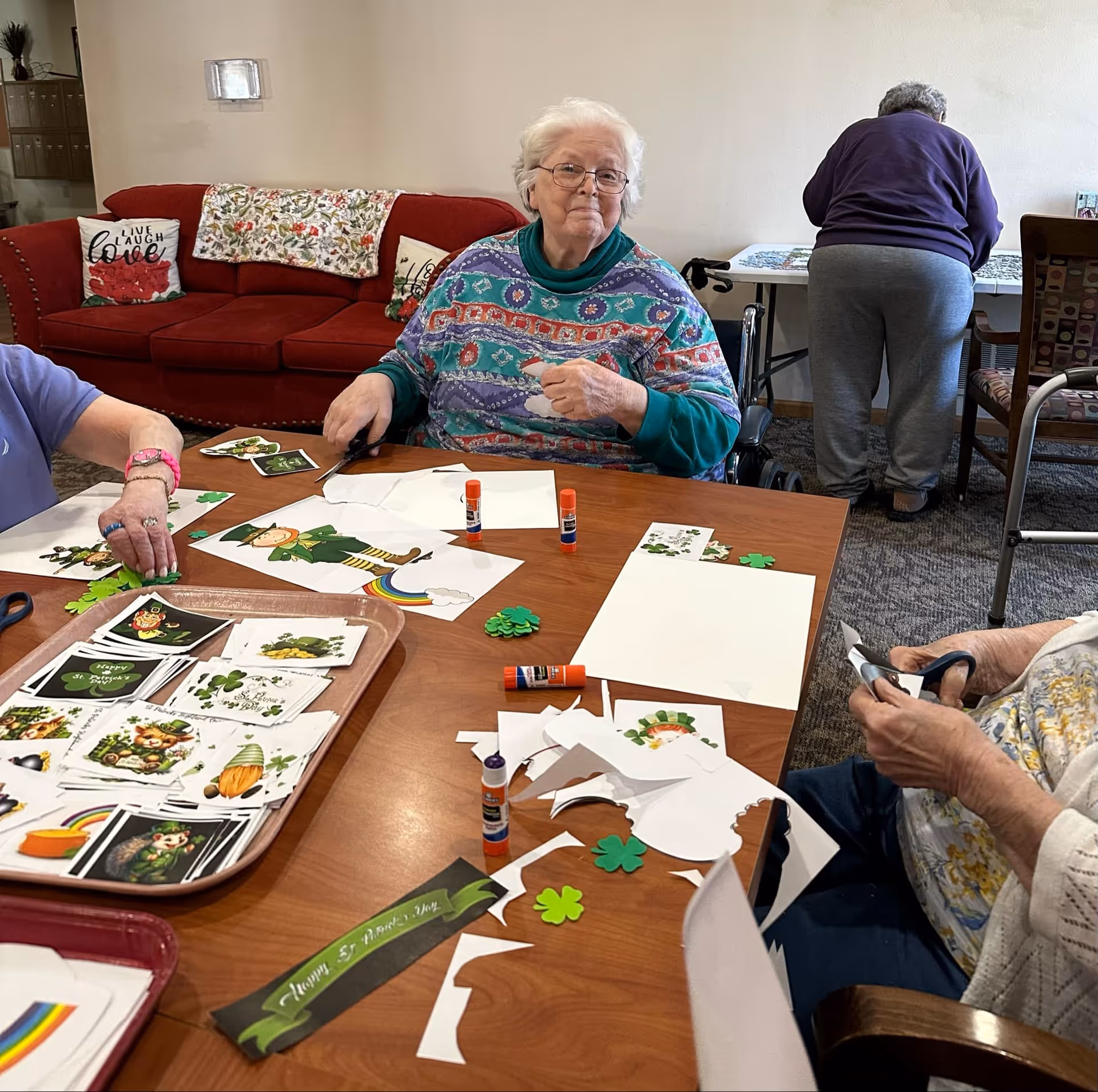 Several elderly individuals engaged in a craft activity at a wooden table, cutting and gluing St. Patrick's Day themed decorations such as shamrocks and leprechauns. One woman in a colorful sweater sits in a wheelchair facing the camera, while another person is standing and working at a table in the background. A red couch with decorative pillows is visible behind them.