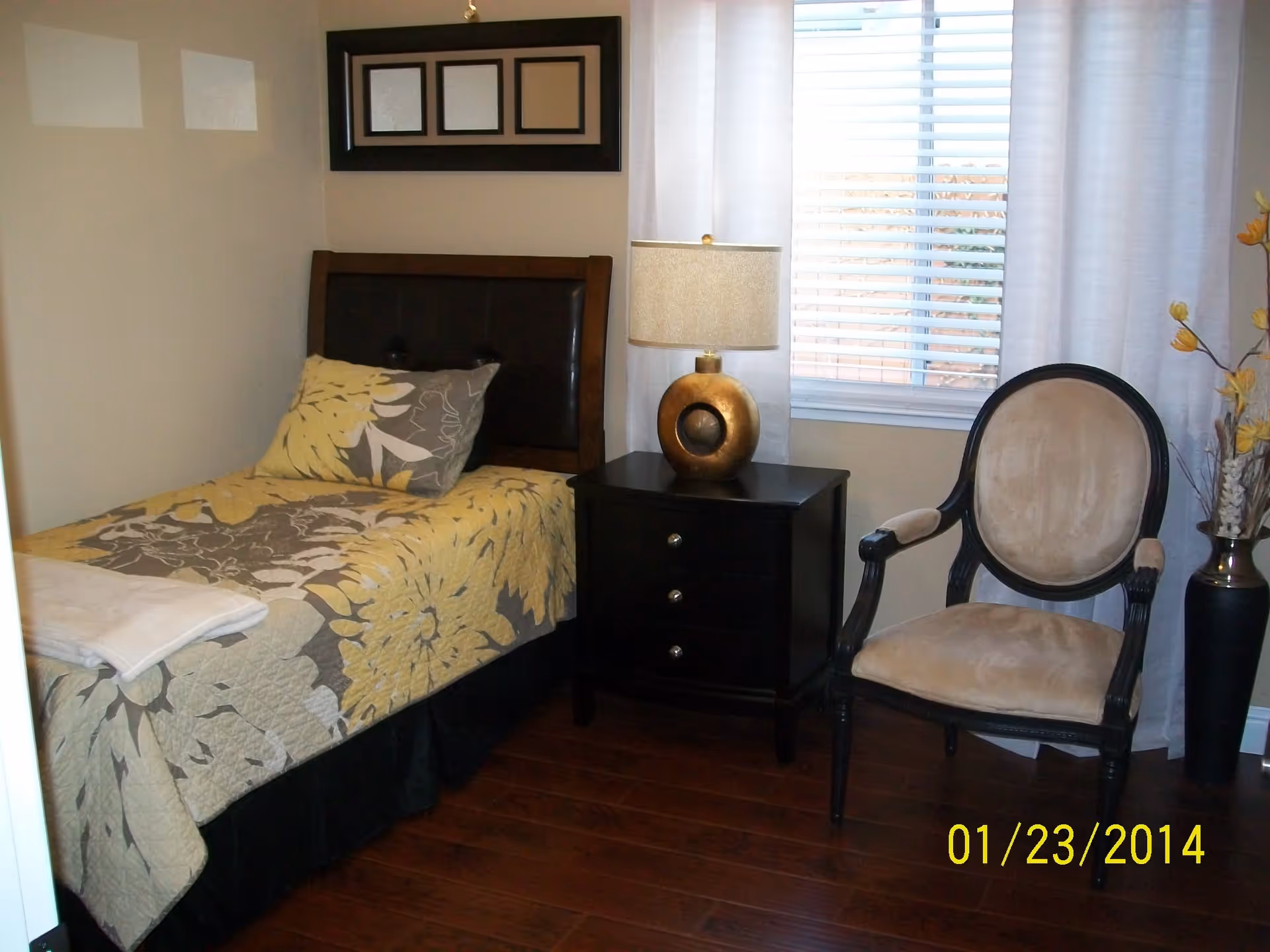 Small furnished bedroom with a single bed, nightstand and lamp next to a window and an upholstered chair.