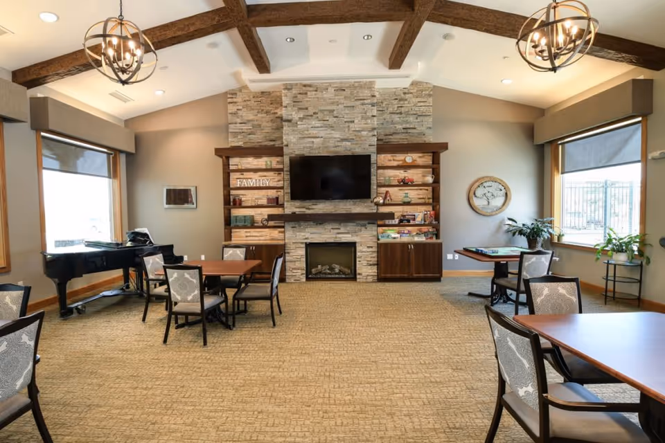 A spacious senior living common area with beige carpet and light gray walls. The room features a stone fireplace with a mounted flat-screen TV above it, flanked by wooden shelves and cabinets. Several tables with chairs are arranged around the room, and a black grand piano is positioned near a large window on the left. Two large windows on either side let in natural light, and there are decorative plants and wall art. The ceiling has exposed wooden beams and two spherical chandeliers.