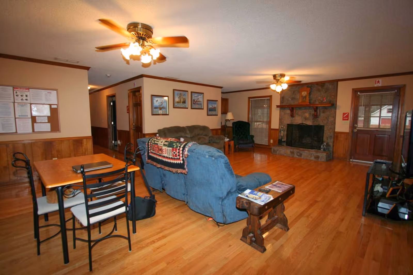 A cozy living room area with wooden flooring and wood-paneled walls. The room features a blue recliner sofa with a colorful blanket draped over the back, a green armchair, and a brown sofa. There is a stone fireplace with a wooden mantel, a wooden door with glass panels, and a dining table with four chairs. Ceiling fans with lights are mounted on the ceiling, and framed pictures hang on the walls.