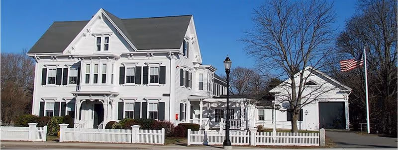 A large white multi-story house with black shutters and a gray roof, surrounded by a white picket fence. There is a street lamp and an American flag on a flagpole in front of the house. Trees without leaves are visible in the background under a clear blue sky.