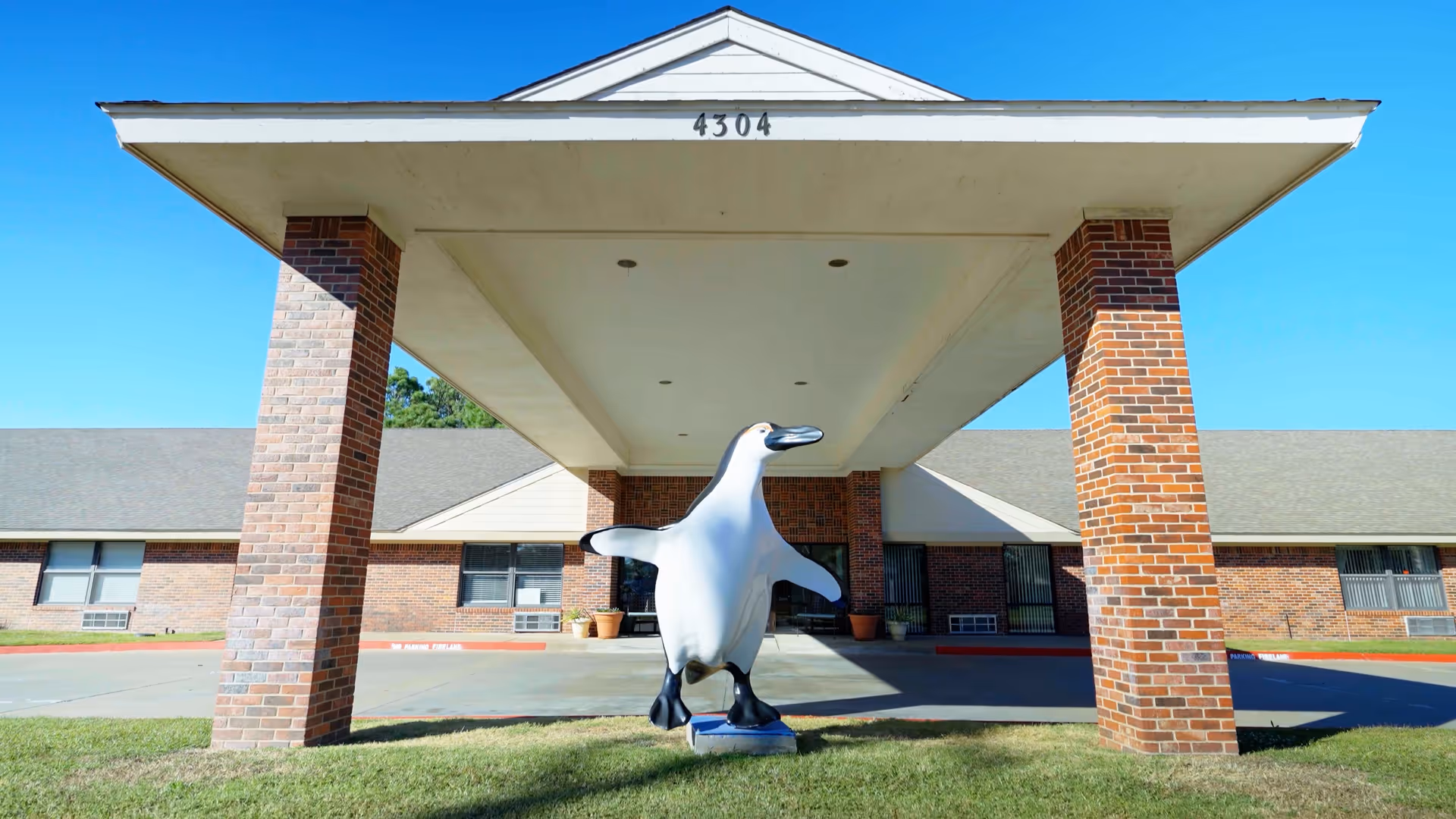 Entrance of a brick building with a covered driveway supported by four brick pillars. A large penguin statue is positioned on the grass under the covered area. The building has several windows and a gray roof, with the number 4304 displayed on the front edge of the roof.