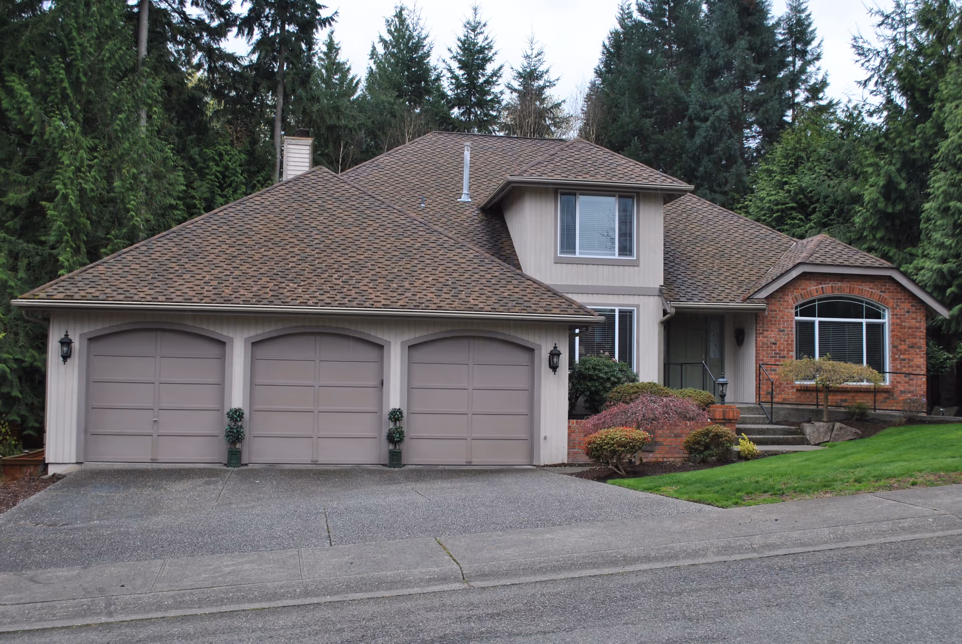 Front exterior view of a single-family house with a three-car garage, beige siding, brick accents, and a well-maintained lawn and shrubs, surrounded by tall evergreen trees.