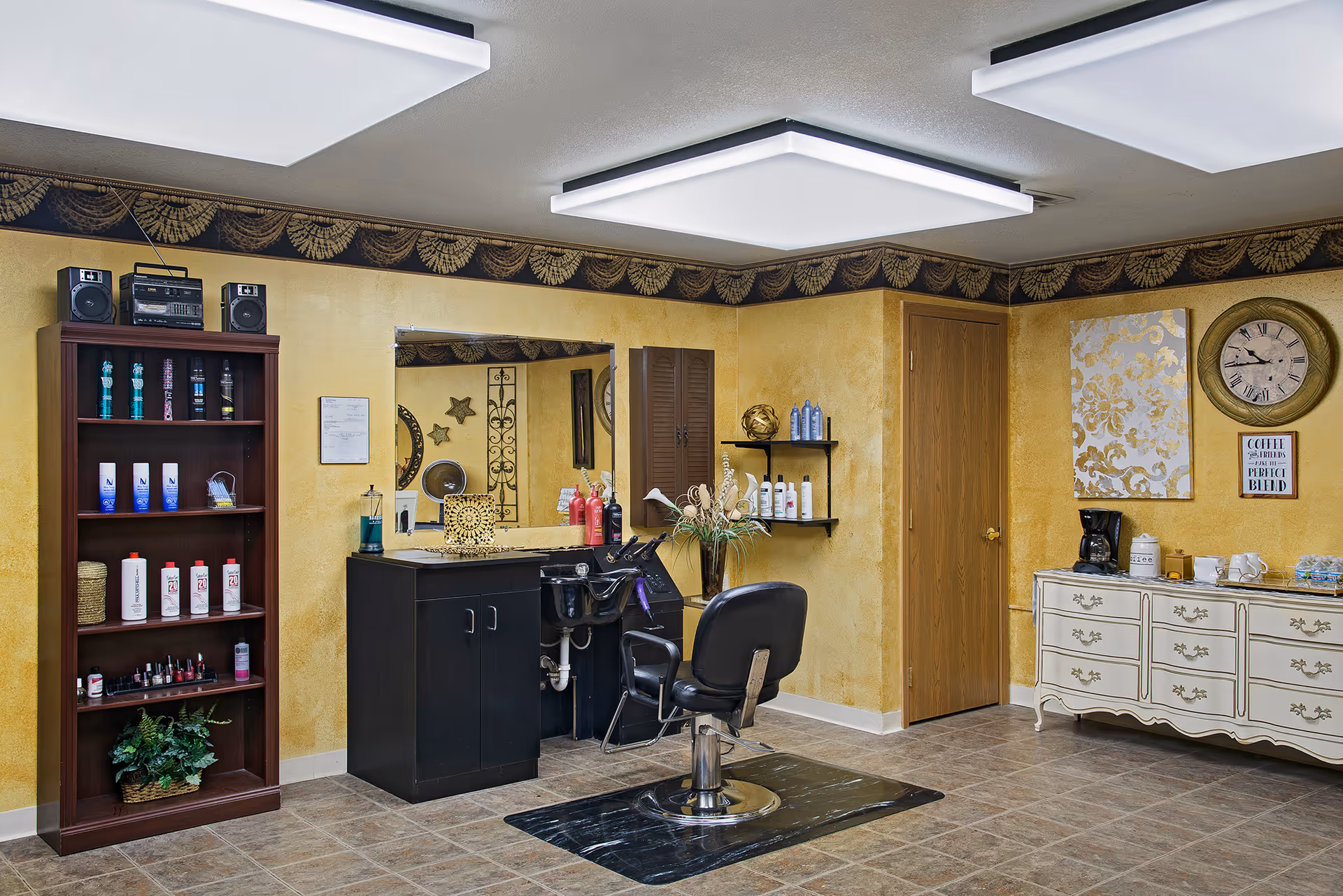 Well-lit interior salon area with a styling chair and sink in front of a mirror, shelves of hair products, and a dresser with a coffee station.