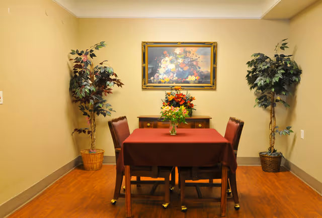 A small dining area with a square table covered by a burgundy tablecloth, surrounded by four wooden chairs with brown cushions. A vase with flowers is placed on the table. Behind the table is a wooden sideboard with a floral arrangement on top and a framed painting of a fruit and flower still life hanging on the beige wall. Two potted artificial plants are positioned in the corners of the room on a wooden floor.