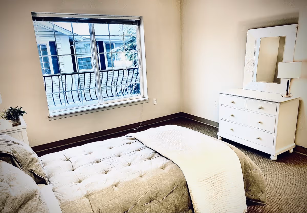 A bright bedroom with a large window showing the exterior of another building. The room features a bed with a tufted beige comforter and pillows, a white dresser with a mirror, and a small lamp on top. There is also a small plant on a nightstand beside the bed.