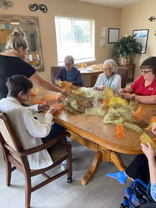 A group of elderly women sitting around a wooden table in a well-lit room, engaging in a craft activity with colorful mesh ribbons. A caregiver is assisting them. The room has a large window, a mirror on the wall, a plant, and some framed pictures.