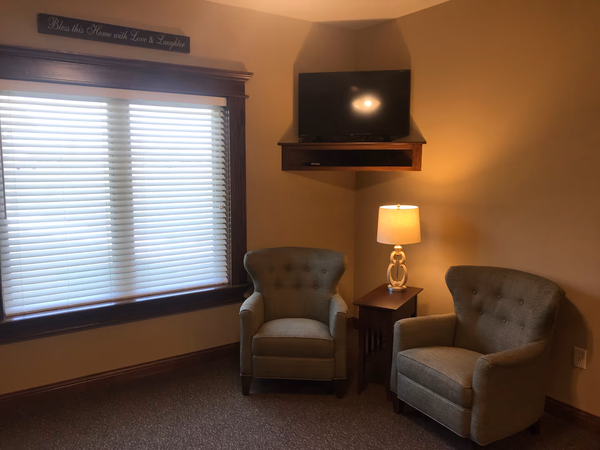 A cozy corner of a living room with two gray upholstered armchairs, a small wooden side table with a lamp, a wall-mounted flat-screen TV above the table, and a large window with white blinds. Above the window is a decorative sign that reads 'Bless this Home with Love & Laughter.'