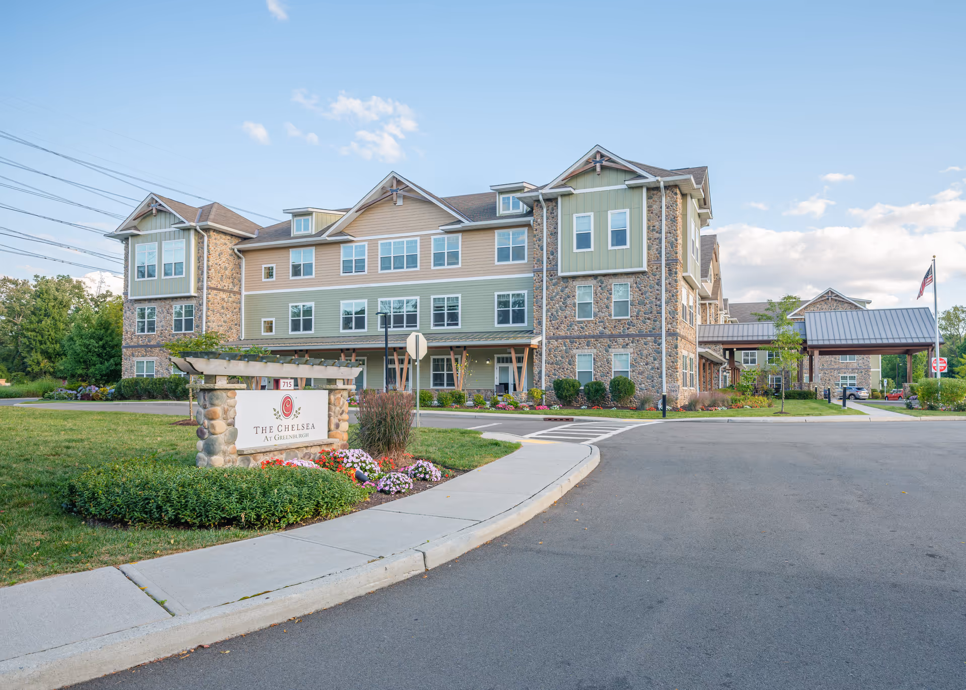 Exterior view of The Chelsea at Greenburgh, a multi-story senior living facility with stone and siding facade, surrounded by landscaped greenery and a clear blue sky.