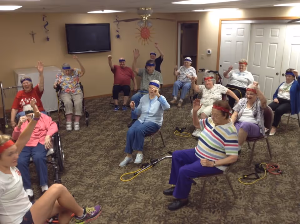 A group of elderly people seated in a room participating in a seated exercise class led by an instructor. They are wearing colorful headbands and raising one arm, following the instructor's movements. The room has carpeted flooring, a wall-mounted TV, and some wall decorations including a sun and stars.