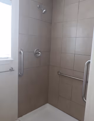Tile-lined shower stall with a wall-mounted showerhead and stainless steel grab bars, plus a small window at the side.