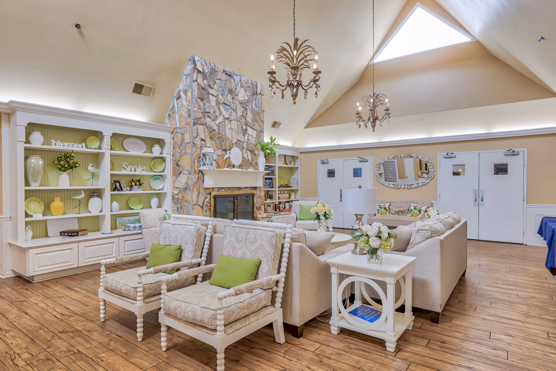 A bright and spacious living room with a high vaulted ceiling and skylight. The room features a stone fireplace in the center, flanked by built-in shelves decorated with vases, plates, and decorative items. There are two cushioned armchairs with green pillows, a beige sectional sofa with floral pillows, and white side tables with flower arrangements. Two ornate chandeliers hang from the ceiling, and double doors are visible in the background.