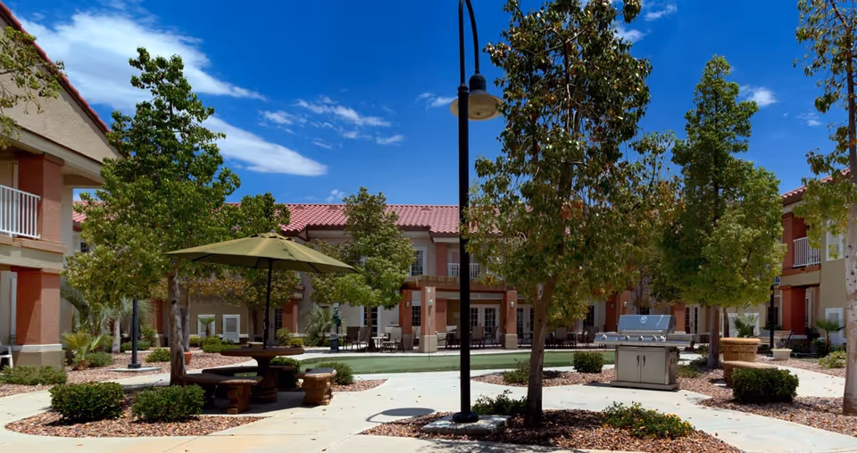 Outdoor courtyard area of a senior living facility with trees, bushes, a green umbrella shading a round picnic table with benches, a barbecue grill, and surrounding two-story buildings under a blue sky with some clouds.
