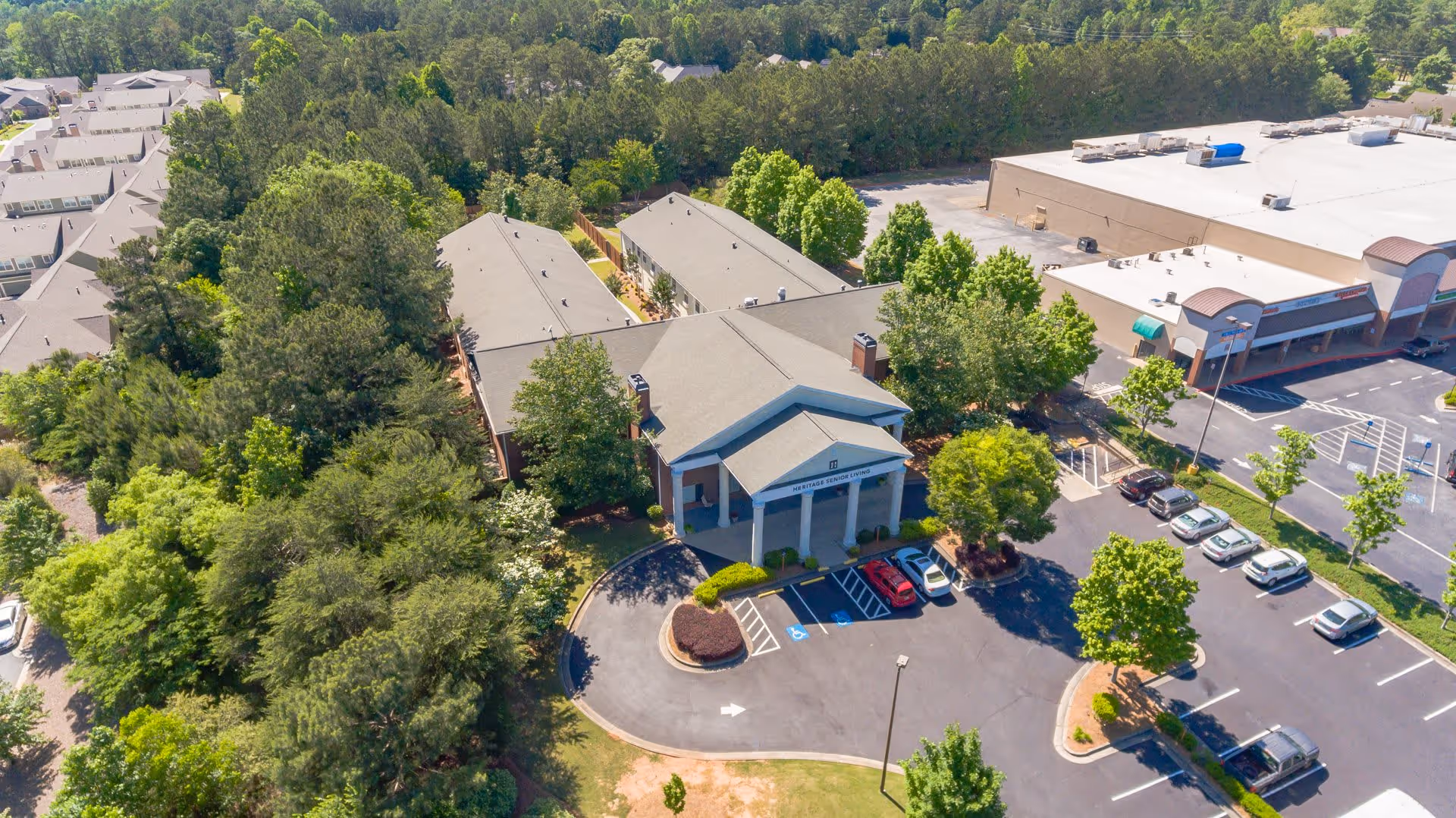 Aerial view of Serenity Empire at Marietta facility surrounded by trees and greenery. The building has a large entrance with columns and a parking lot with several cars. Adjacent to the facility is a large commercial building with a white roof and parking spaces.
