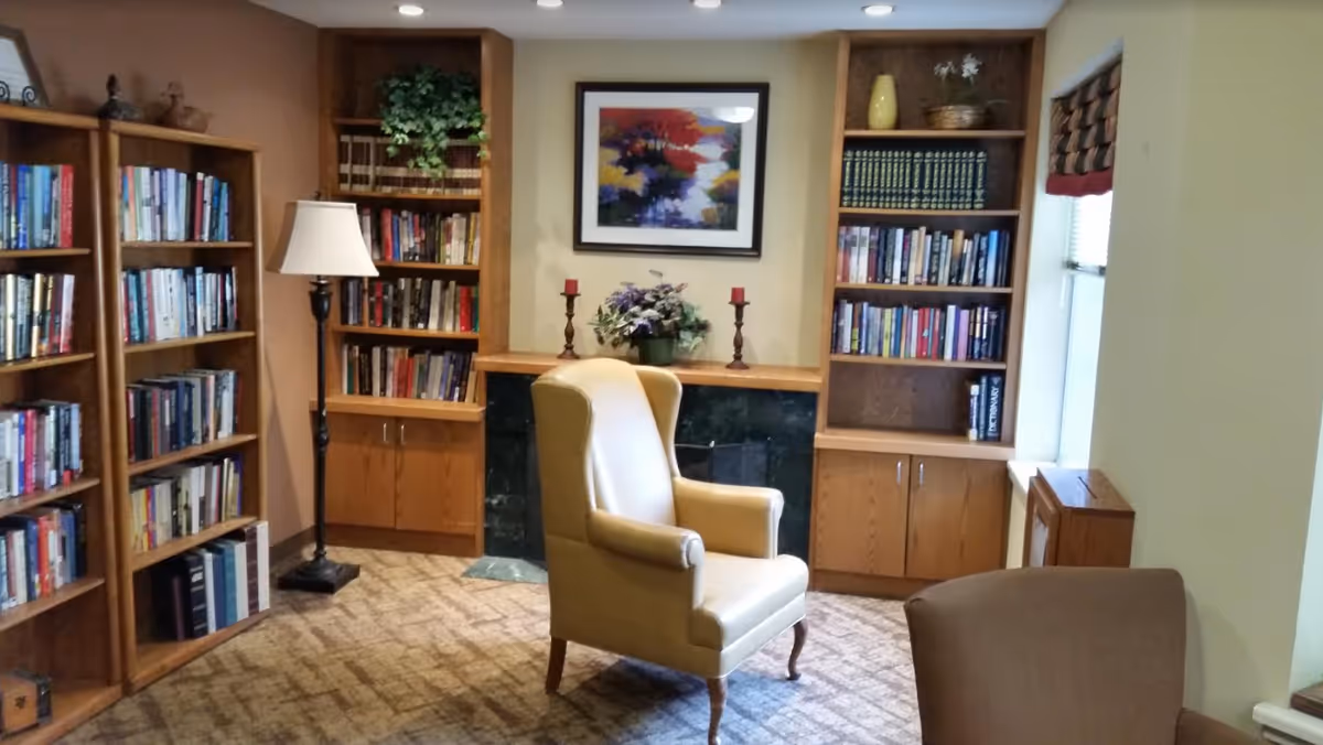 A cozy reading room with wooden bookshelves filled with books, a beige leather armchair in the center, a floor lamp, a framed colorful painting on the wall, and decorative plants and candles on a mantelpiece.