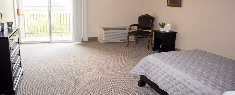 Bedroom in a senior living facility featuring a bed, dresser, chair, nightstand, and sliding balcony door.