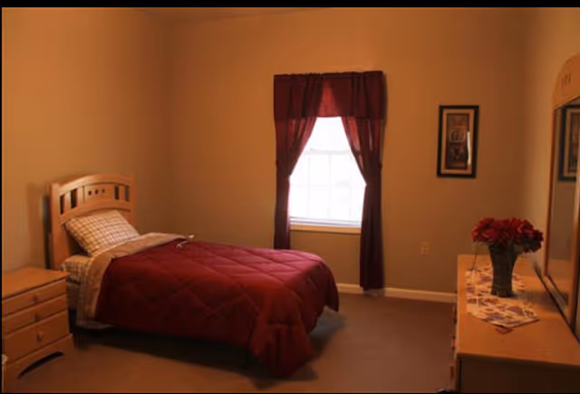 Small bedroom with a single bed dressed in a burgundy comforter, a window with matching curtains, and a dresser topped with a vase of red flowers.