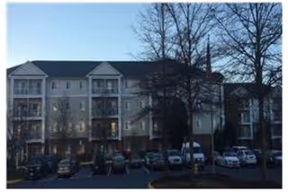 Exterior view of a multi-story senior living facility building with balconies, several parked cars in front, and leafless trees in the foreground during dusk or early evening.