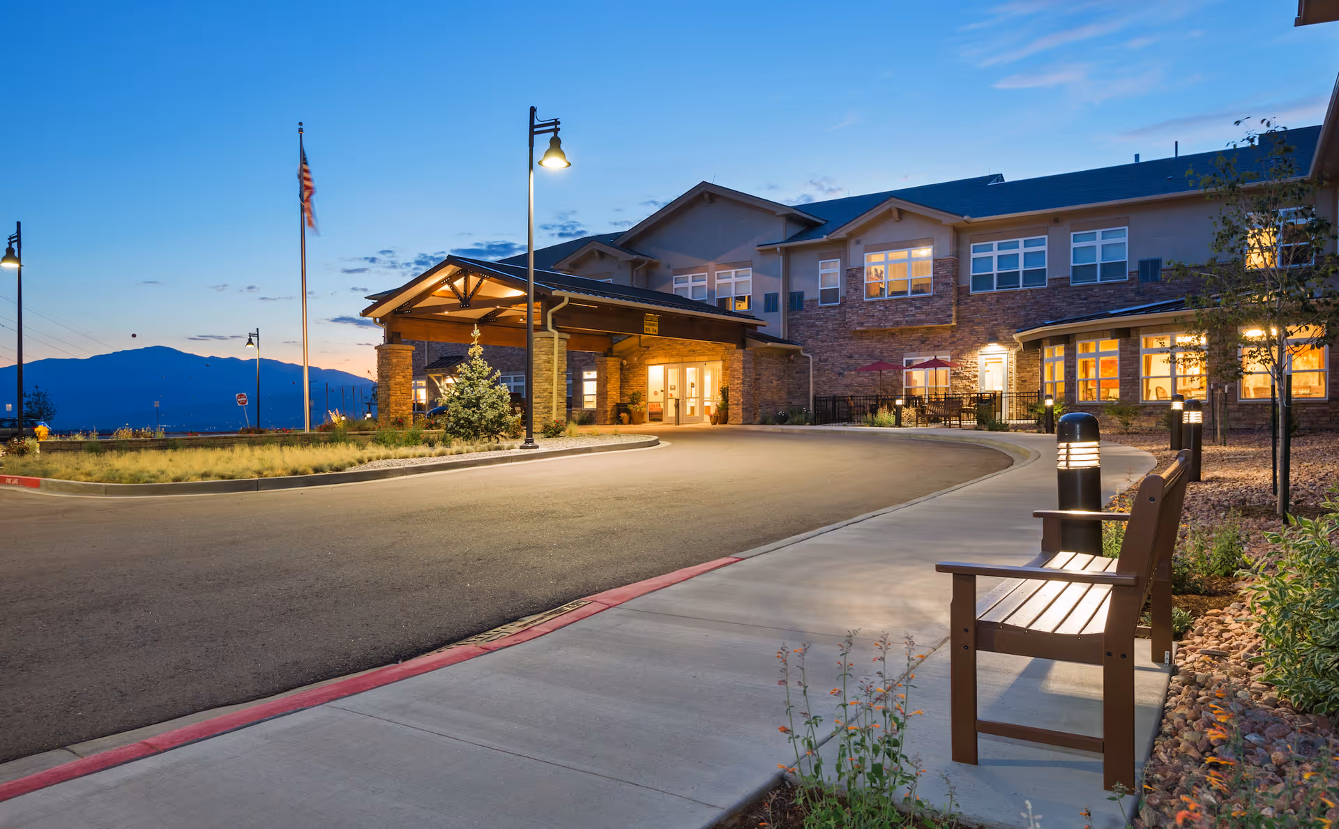 Exterior view of Melody Living facility at dusk with a curved driveway, a covered entrance, lit street lamps, an American flag, and a bench along the sidewalk. The building has multiple windows with warm interior lighting and a stone facade.