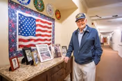 An elderly man wearing a cap and jacket stands smiling in a well-lit hallway of a senior living facility. Behind him on the wall is a large American flag quilt surrounded by military service emblems. On a countertop below the quilt are framed photographs and memorabilia.