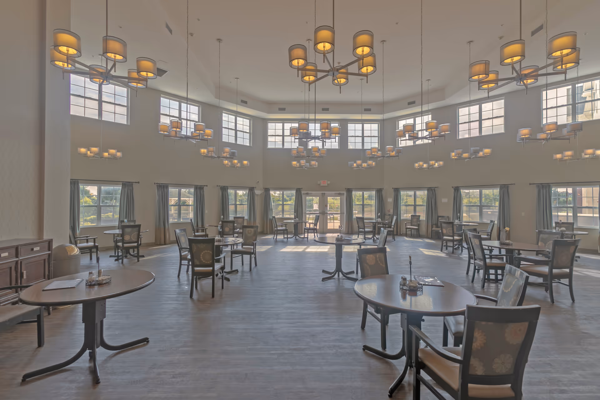 Spacious dining room with multiple round tables and chairs arranged neatly. Large windows with curtains allow natural light to fill the room. Several modern chandeliers hang from the high ceiling, illuminating the space.