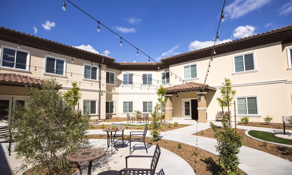 Outdoor courtyard area of The Pointe At Summit Hills facility with tables and chairs, string lights overhead, small trees and plants, and a two-story beige building with multiple windows surrounding the courtyard under a blue sky with some clouds.